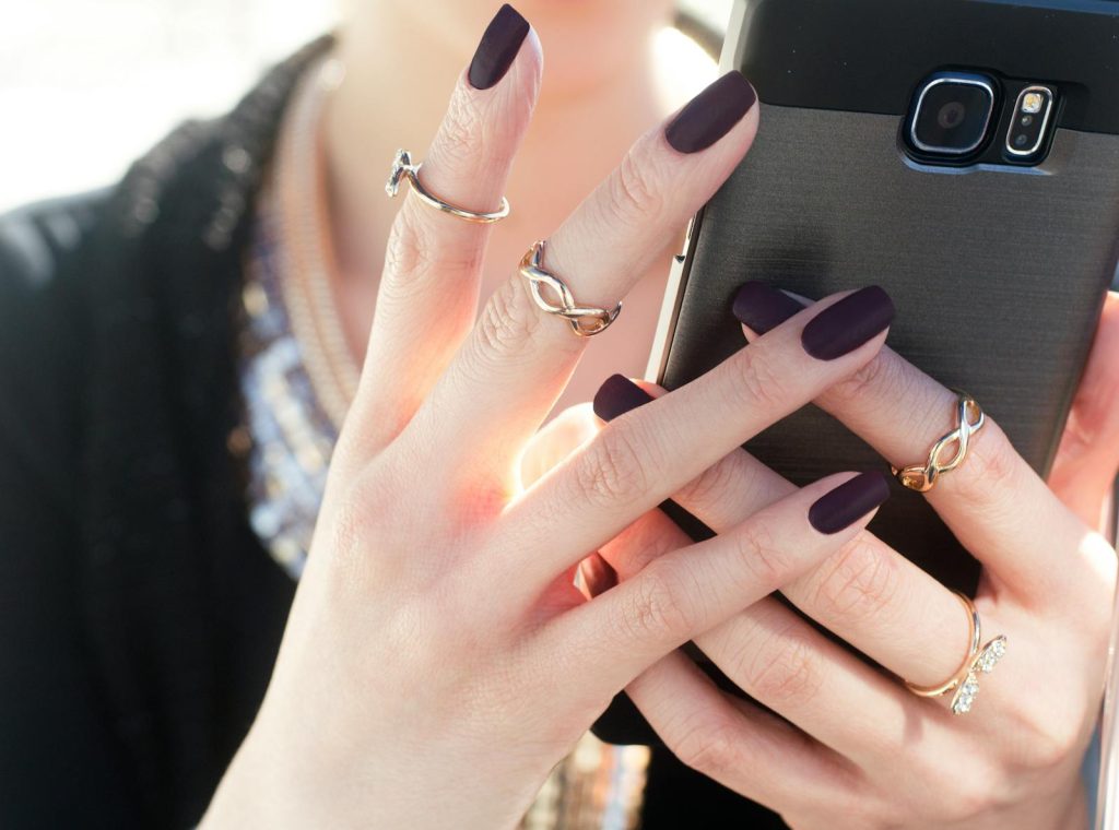 Close-up of a woman with manicured nails and rings holding a smartphone, showcasing fashion elegance.